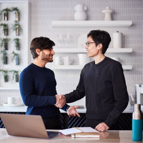 Two people shaking hands beside a desk with a laptop