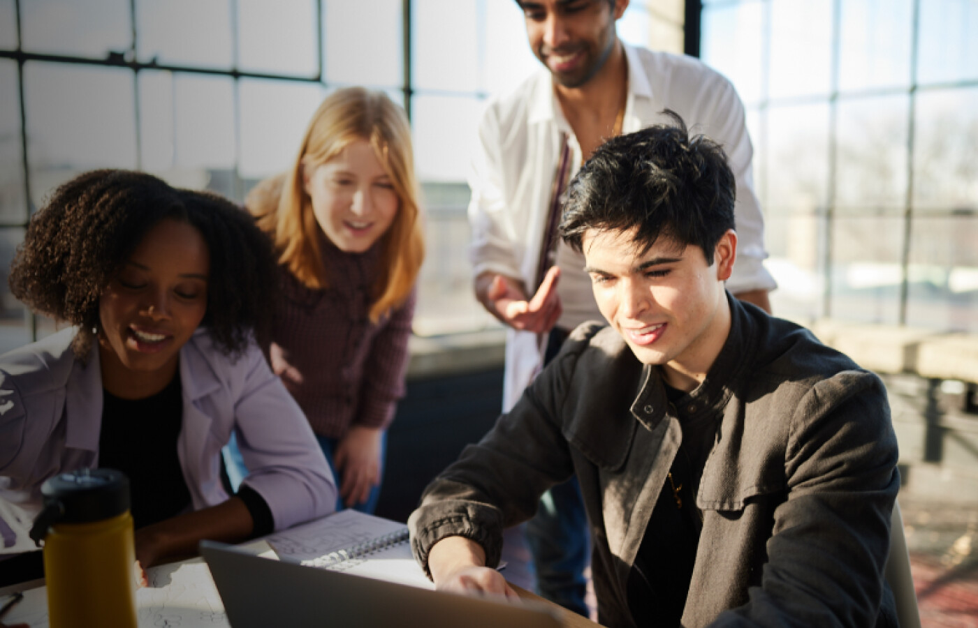 Group of Arts students in a loft standing around a table with a laptop