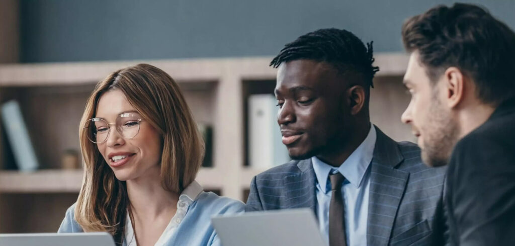 a woman and 2 men in an office working in a meeting with laptops.